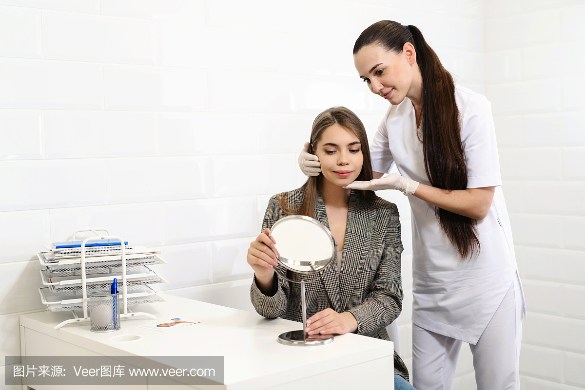 美容门诊会诊。女美容医生与病人交谈,并显示年轻女孩的变化,她的脸使用镜子。规划美容